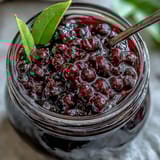 Homemade black currant jam in a glass jar, deep purple and glossy, with fresh blackcurrants scattered around on a rustic wooden table.