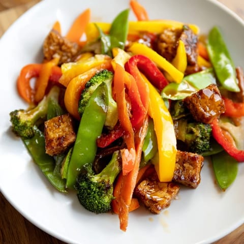 A close-up of Vegetable Tempeh Stir-Fry highlights crisp sugar snap peas, julienned carrots, and aromatic garlic in a savory glaze.